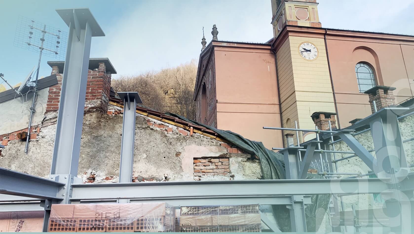 Struttura in acciaio in fase di costruzione davanti a un edificio storico, con pareti in pietra e mattoni parzialmente demolite. Sullo sfondo, una chiesa con torre dell’orologio e facciata color pesca. Presenti impalcature e materiali da cantiere.