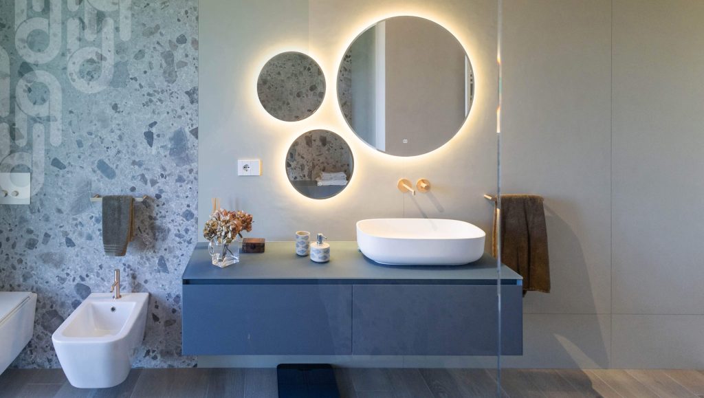 Bagno moderno con mobile lavabo sospeso blu, lavabo ovale bianco e tre specchi rotondi retroilluminati sulla parete; a sinistra bidet e parete con rivestimento effetto terrazzo, accessori decorativi sul piano e asciugamani appesi.