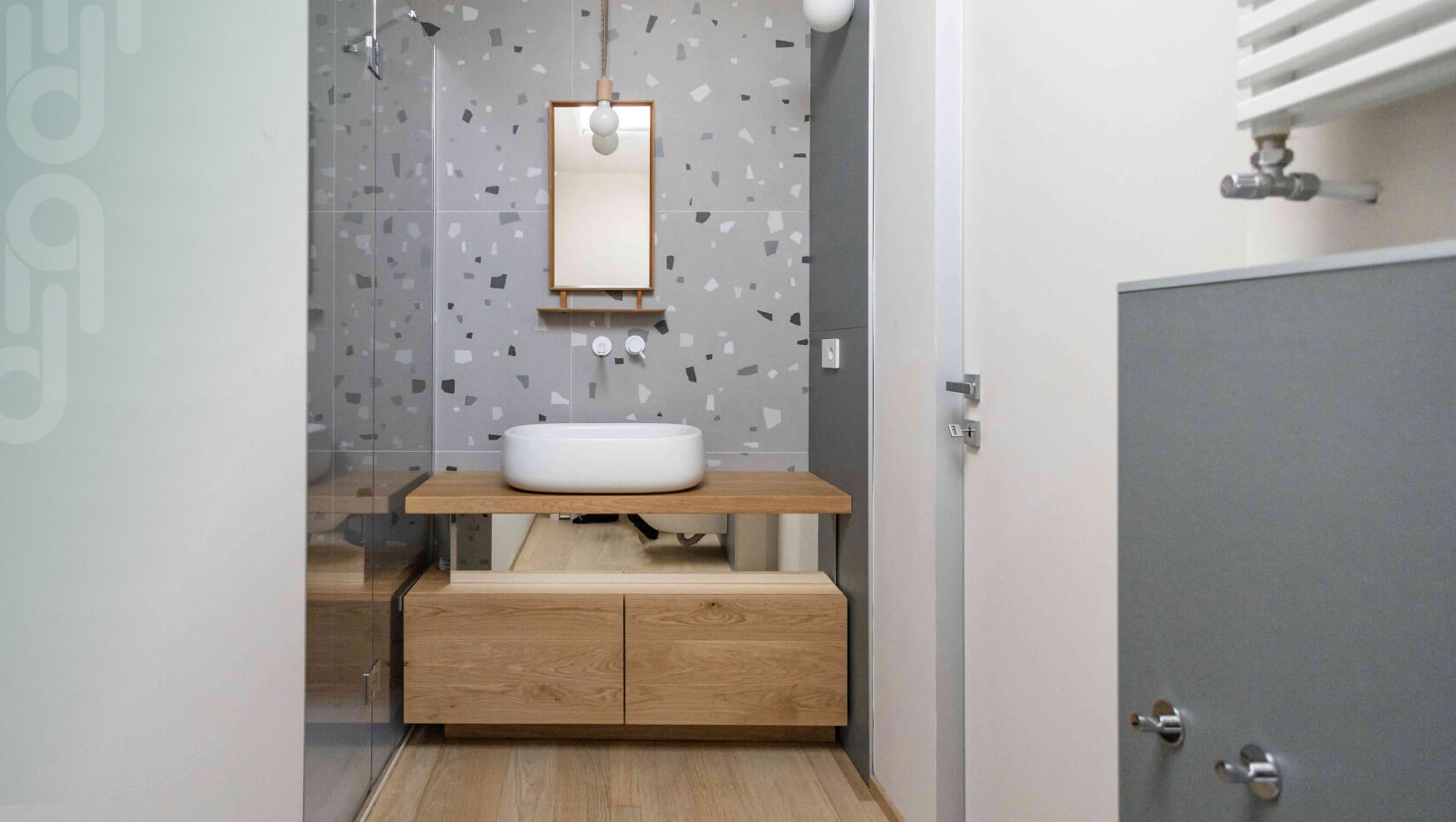 Arredo bagno piccolo e moderno con lavabo da appoggio, mobile sospeso in legno e rivestimento doccia effetto terrazzo veneziano grigio.